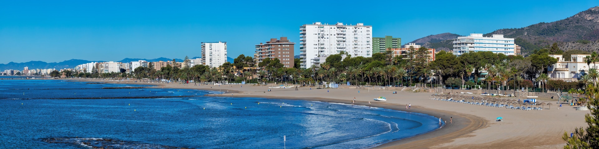 A coastal cityscape with a sandy beach, a rocky breakwater, and a backdrop of mountains.