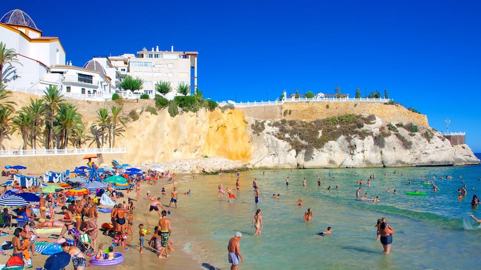 A crowded beach with people swimming and sunbathing, palm trees, and buildings in the background.