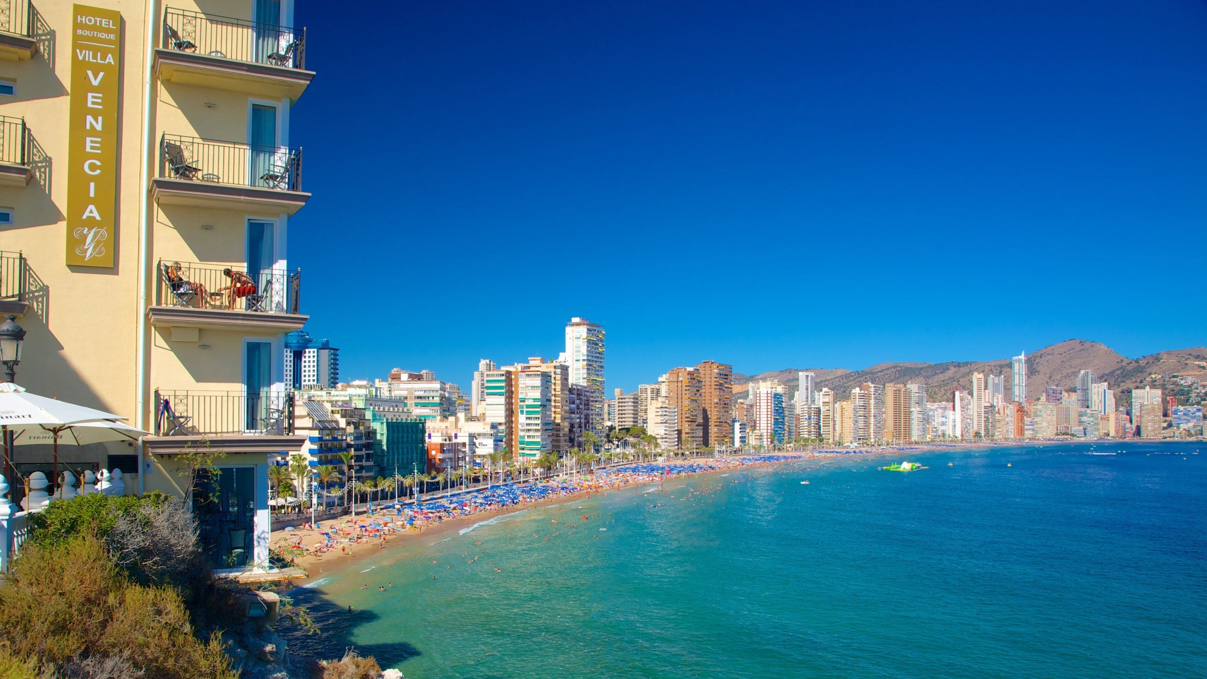 A coastal cityscape with a prominent hotel named "Villa Venezia" on the left, a bustling beach with numerous umbrellas and people, and a clear blue sky.