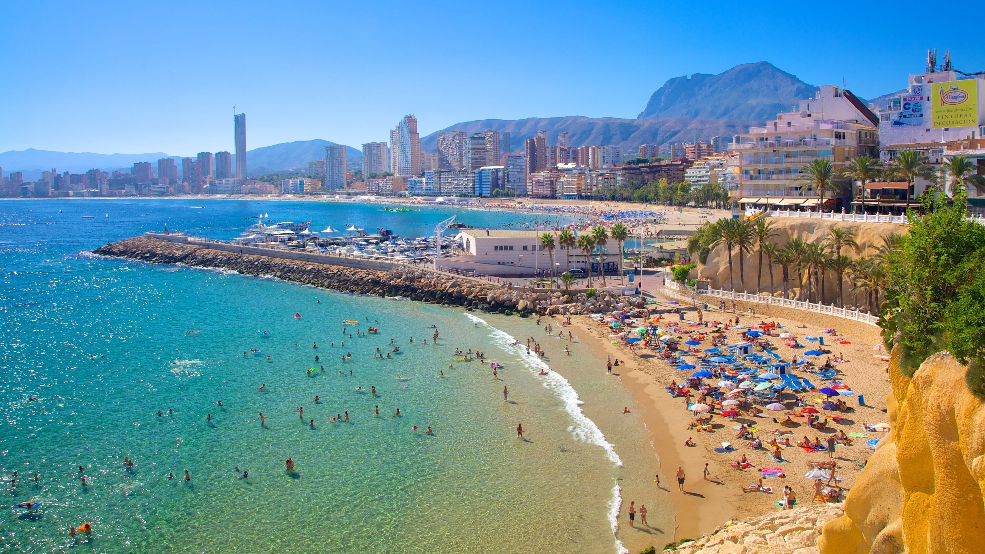 Uma praia cheia com águas turquesa cristalinas, um paredão rochoso e um horizonte urbano com edifícios altos e montanhas ao fundo.