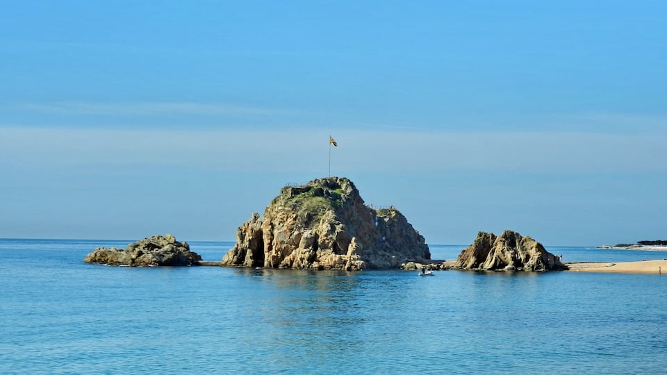 A rocky island with a flag in the center, surrounded by calm blue waters and a clear sky.