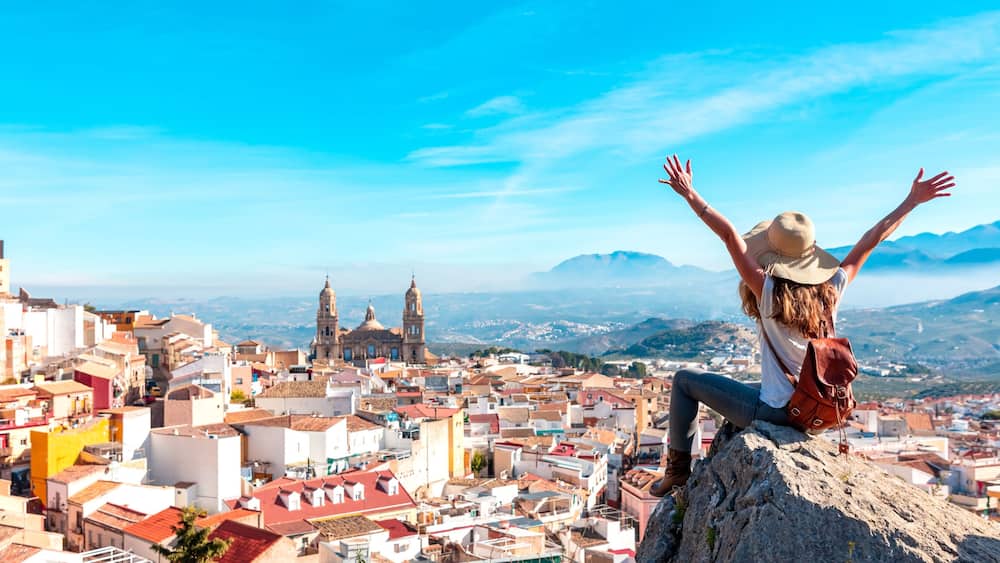 Eine Person steht auf einem Felsen, die Arme in den Himmel erhoben, und blickt auf eine Stadtlandschaft mit historischen Gebäuden und Bergen im Hintergrund.