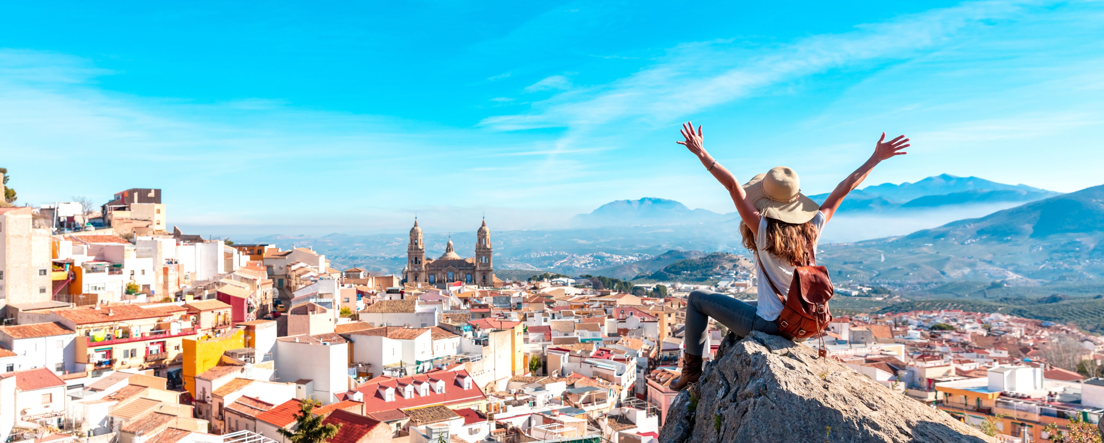 A person on a rock with arms raised, overlooking a cityscape with historic buildings and mountains in the background.