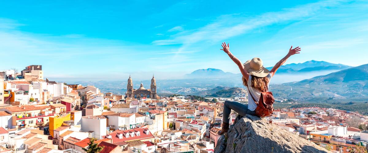 A person on a rock with arms raised, overlooking a cityscape with historic buildings and mountains in the background.