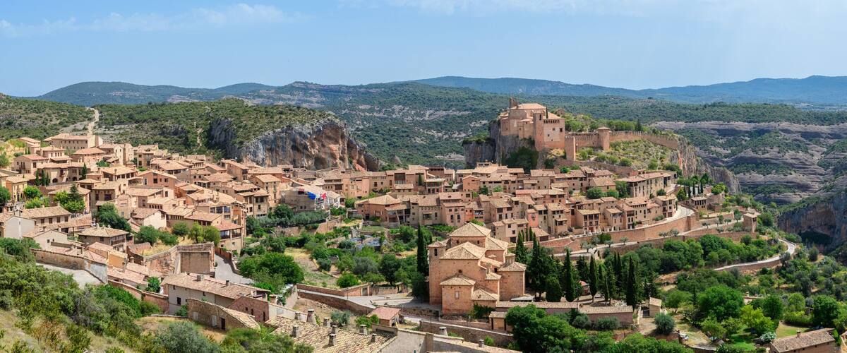 A historic town with terracotta-roof buildings nestled in a valley, surrounded by mountains and greenery.