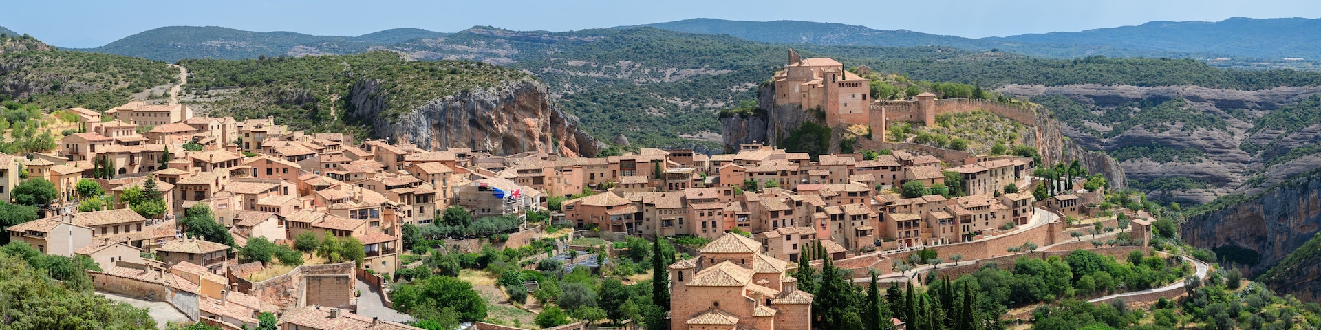 A historic town with terracotta-roof buildings nestled in a valley, surrounded by mountains and greenery.