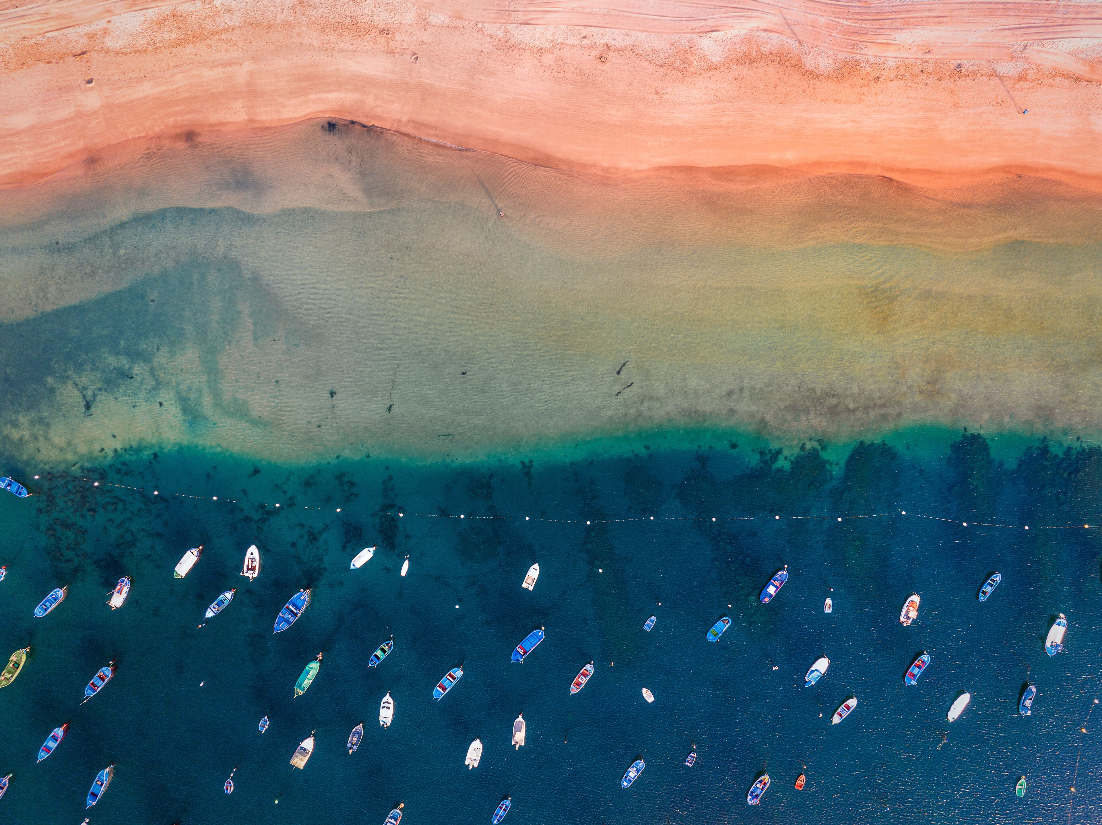 Cena costeira com uma praia arenosa, um degradê claro da água do raso para o fundo, e vários barcos pequenos espalhados pela água.
