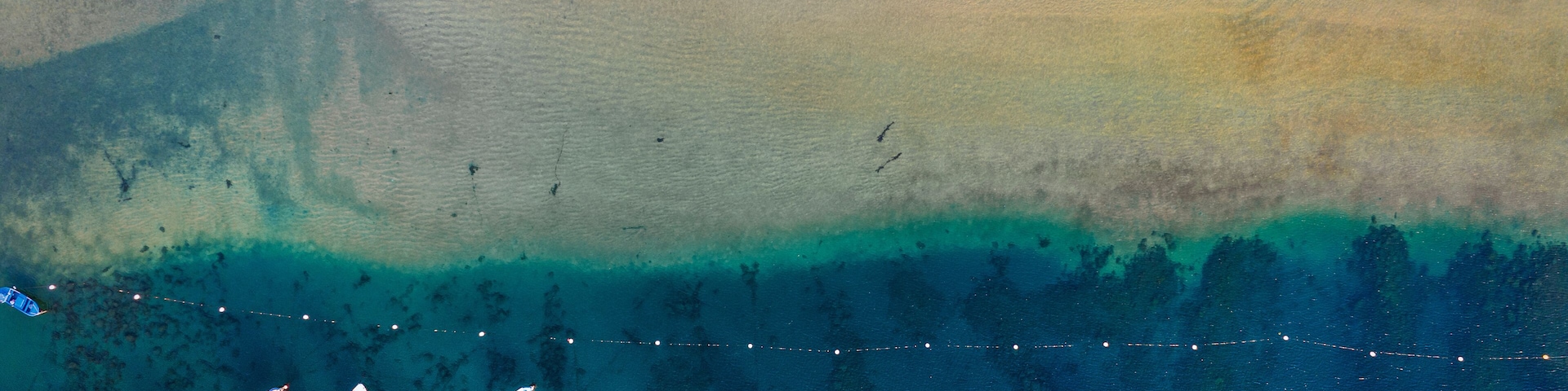 Cena costeira com uma praia arenosa, um degradê claro da água do raso para o fundo, e vários barcos pequenos espalhados pela água.