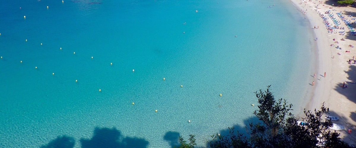 A coastal town with a clear blue bay, a sandy beach, and buildings on a hillside.