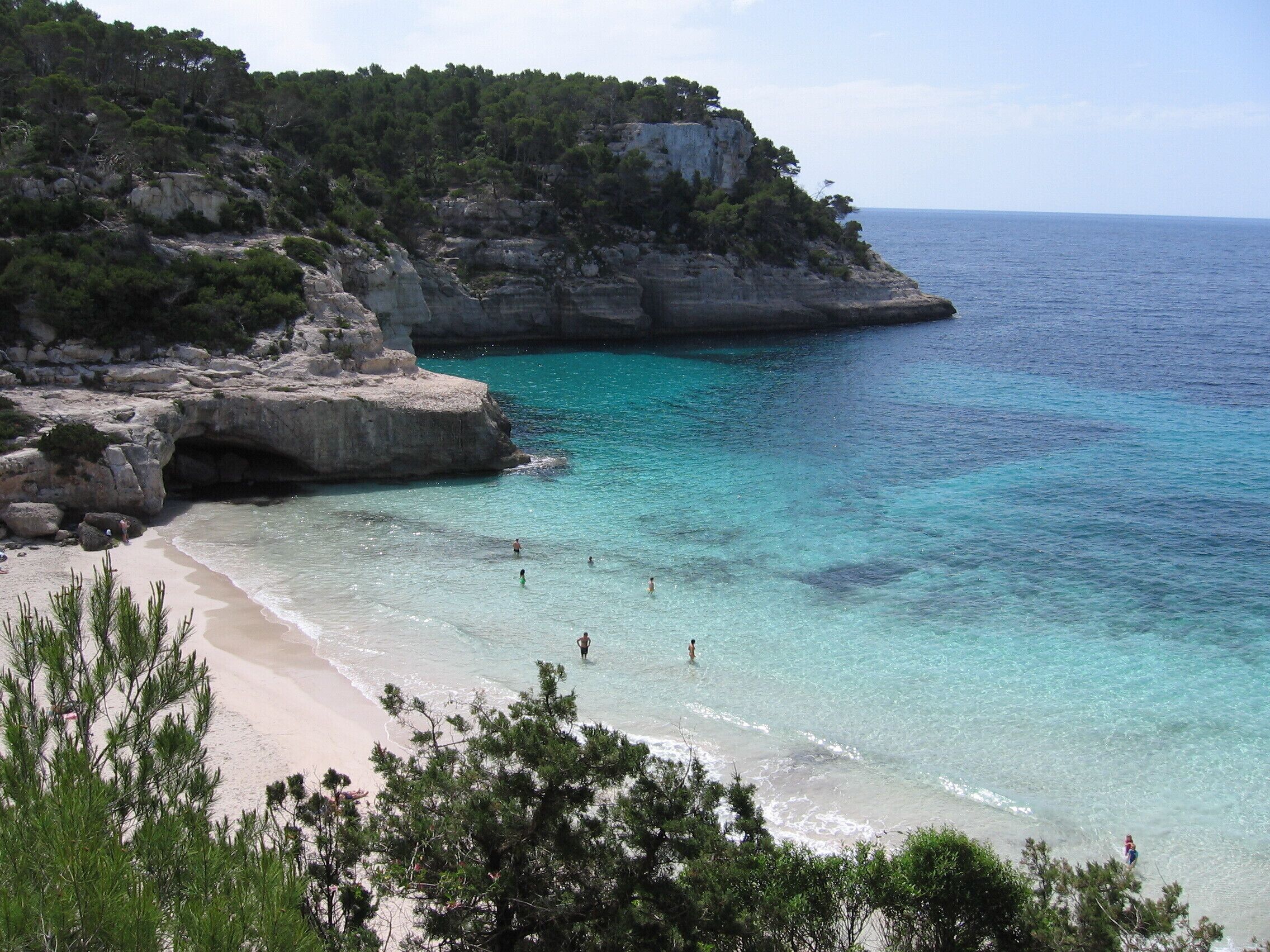 A coastal landscape with clear turquoise waters, a sandy beach, and rocky cliffs with sparse vegetation.
