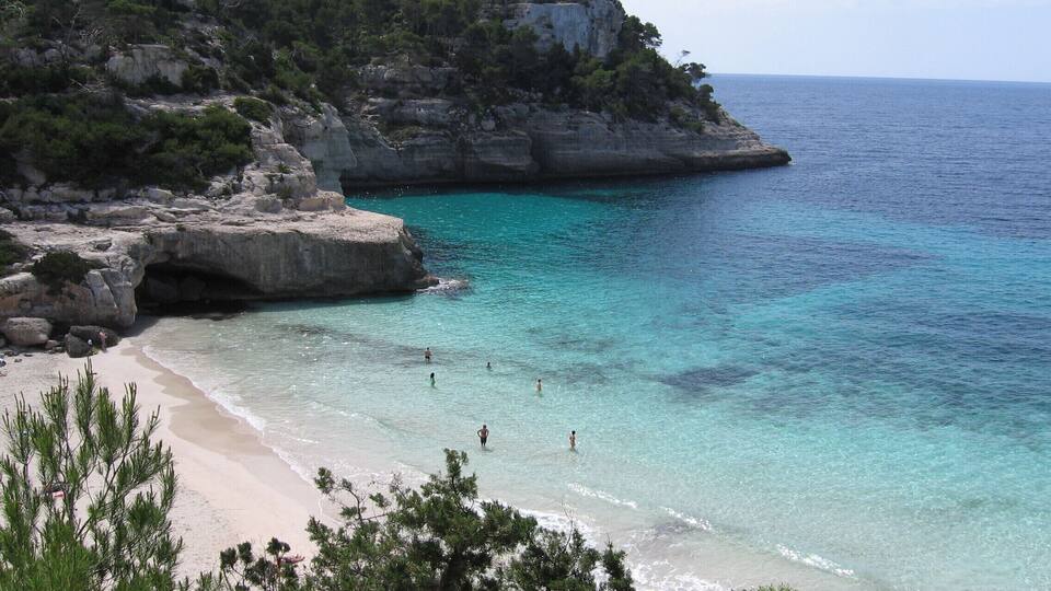 A coastal landscape with clear turquoise waters, a sandy beach, and rocky cliffs with sparse vegetation.