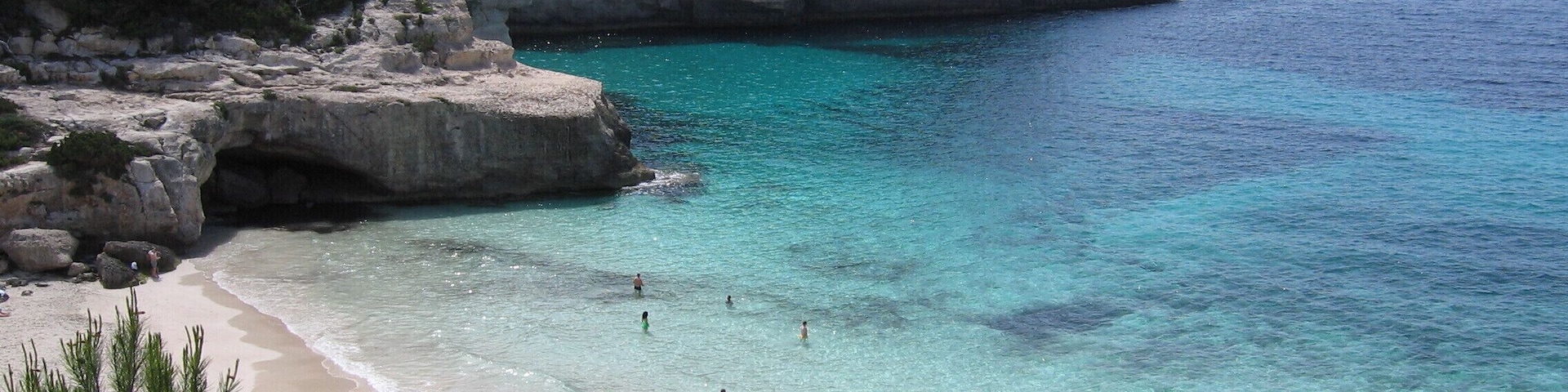 Un paisaje costero con aguas turquesas cristalinas, una playa de arena y acantilados rocosos con vegetación escasa.