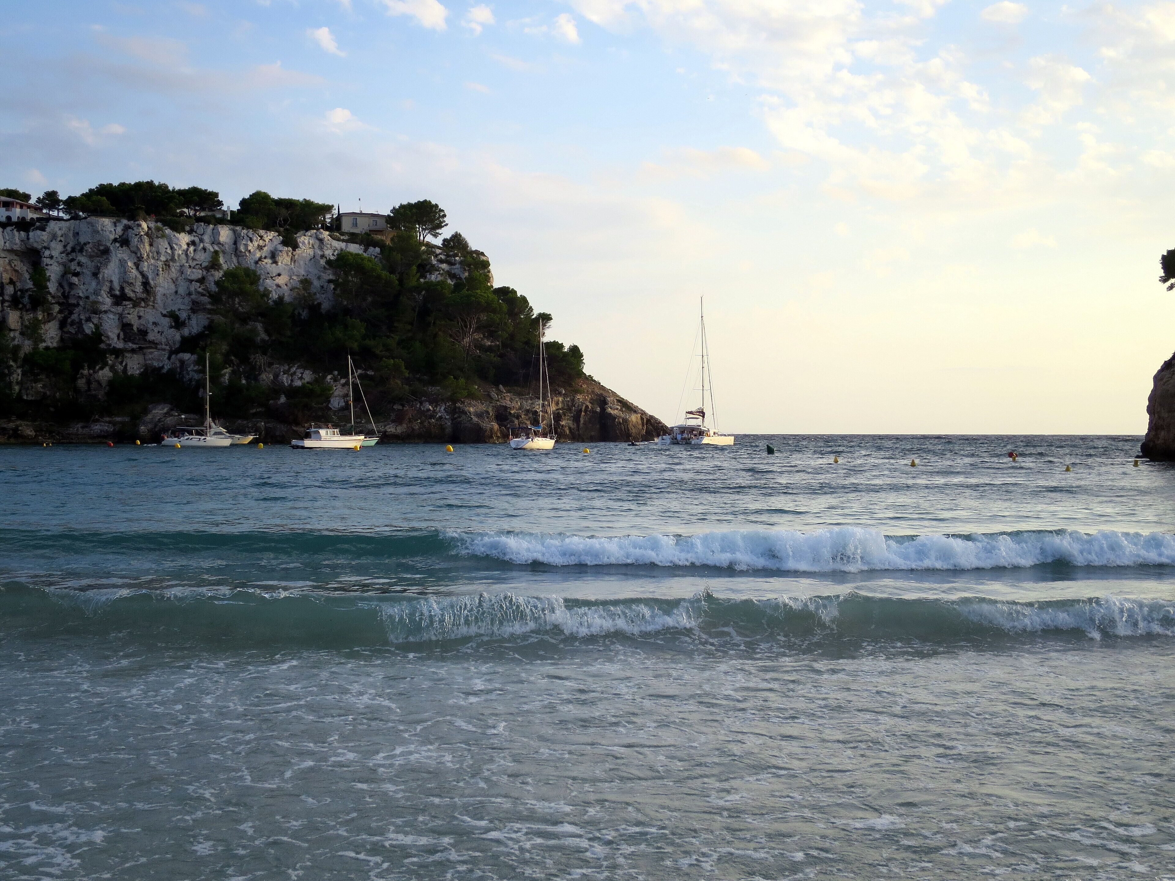 A coastal scene with rocky cliffs, a sailboat, and several yachts anchored near the shore.
