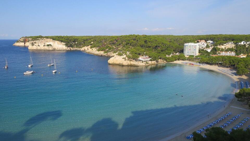 A coastal bay with clear blue waters, sailboats, a sandy beach with umbrellas, and buildings on a hillside.
