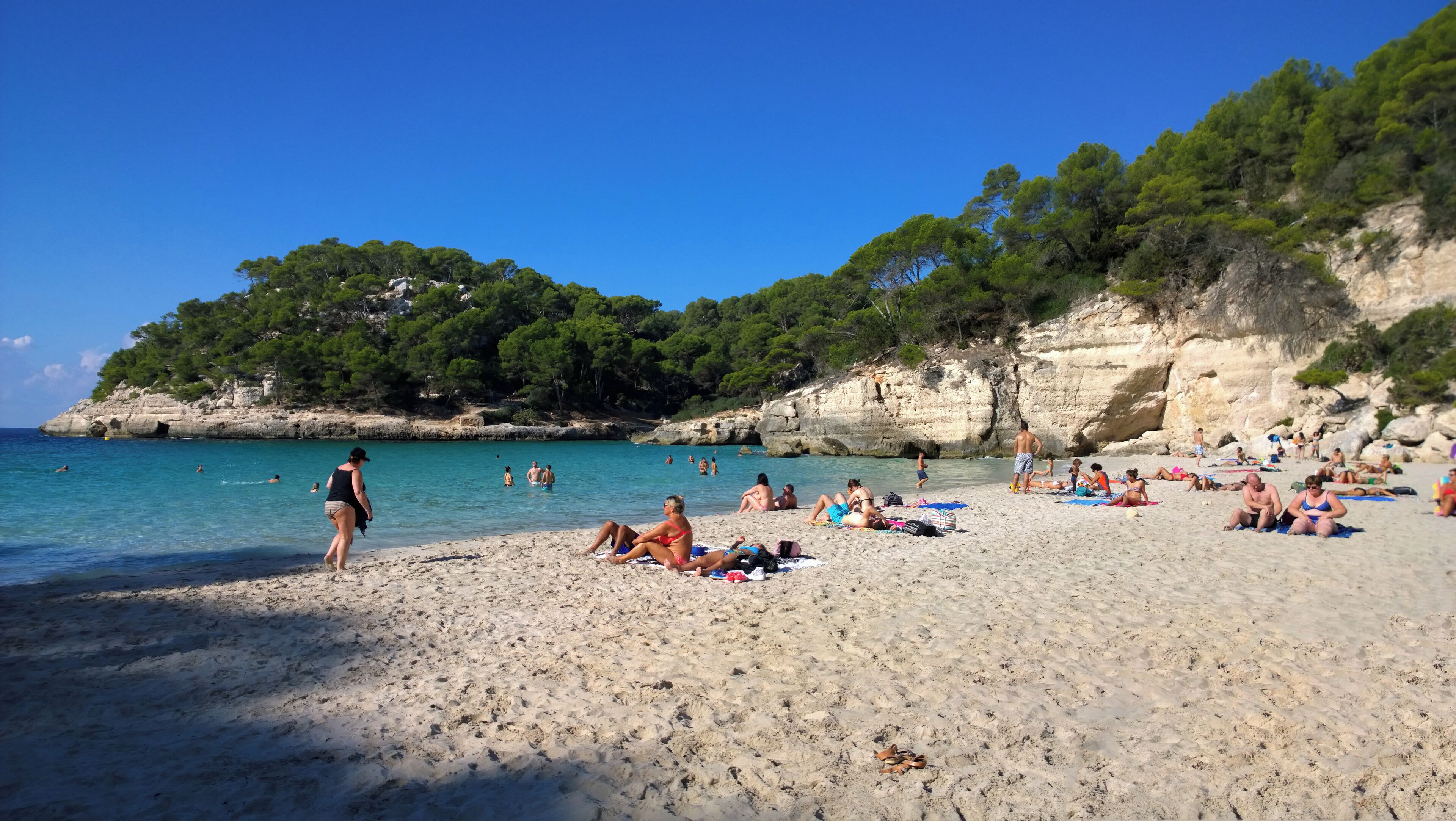 A beach with clear turquoise water, people sunbathing and swimming, and a rocky cliff with greenery in the background.