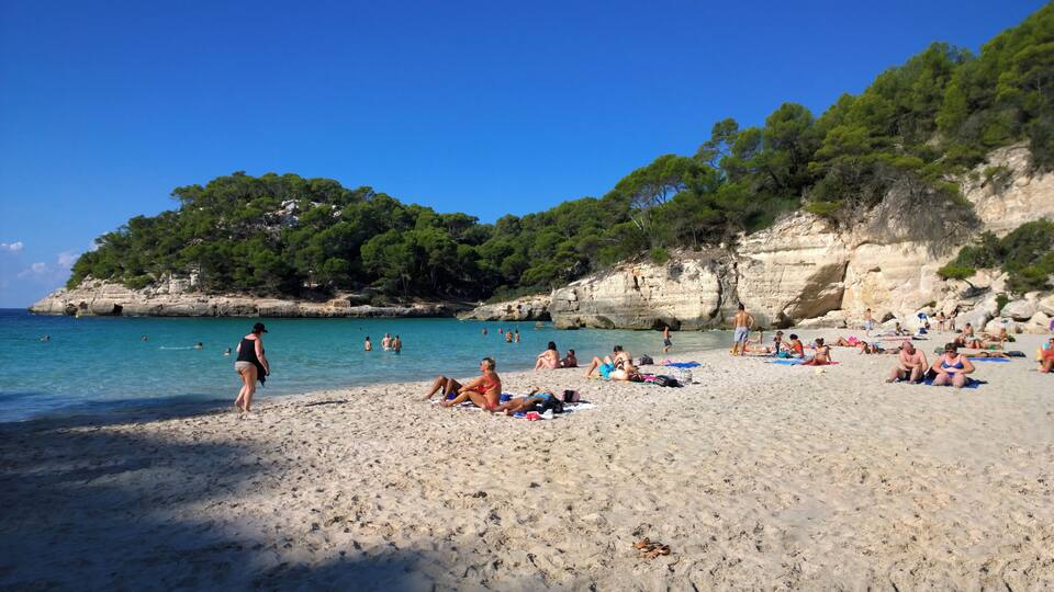 A beach with clear turquoise water, people sunbathing and swimming, and a rocky cliff with greenery in the background.