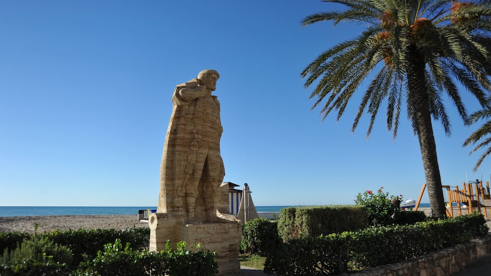 A stone statue, a palm tree, and a beach in the background.