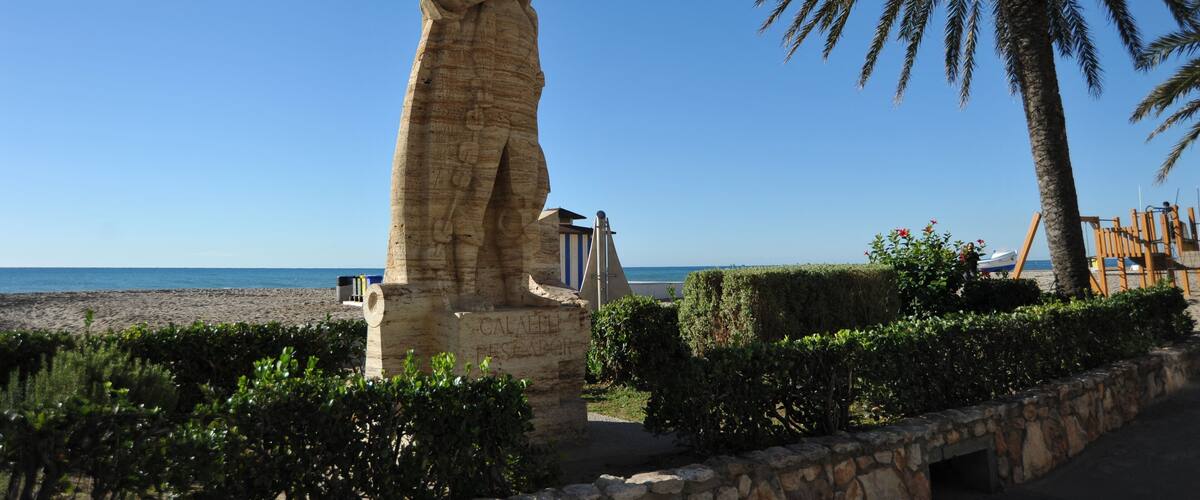 A stone statue, a palm tree, and a beach in the background.