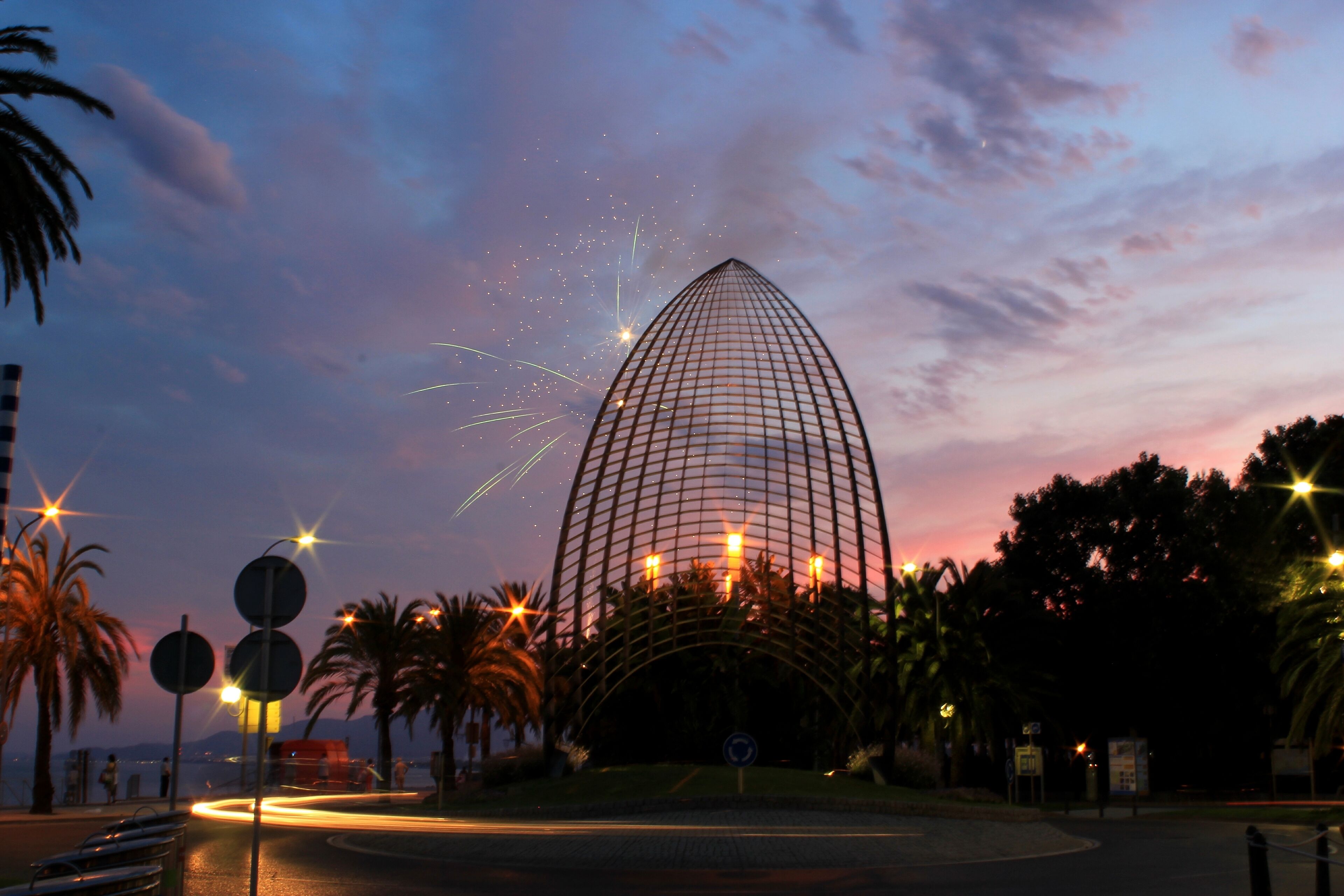 A modern glass building with a curved roof, surrounded by palm trees and street lamps.