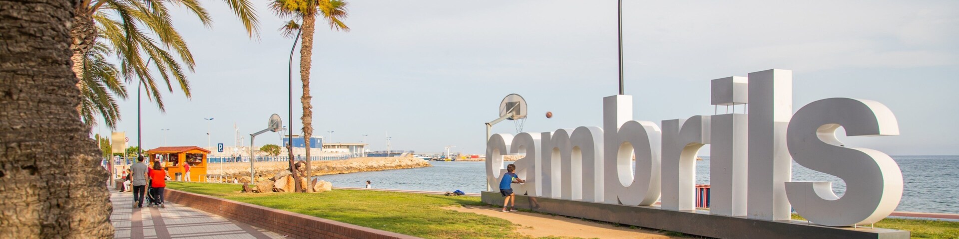 Paseo marítimo costero con palmeras, un gran letrero que dice 'Cambrils' y vistas al mar.