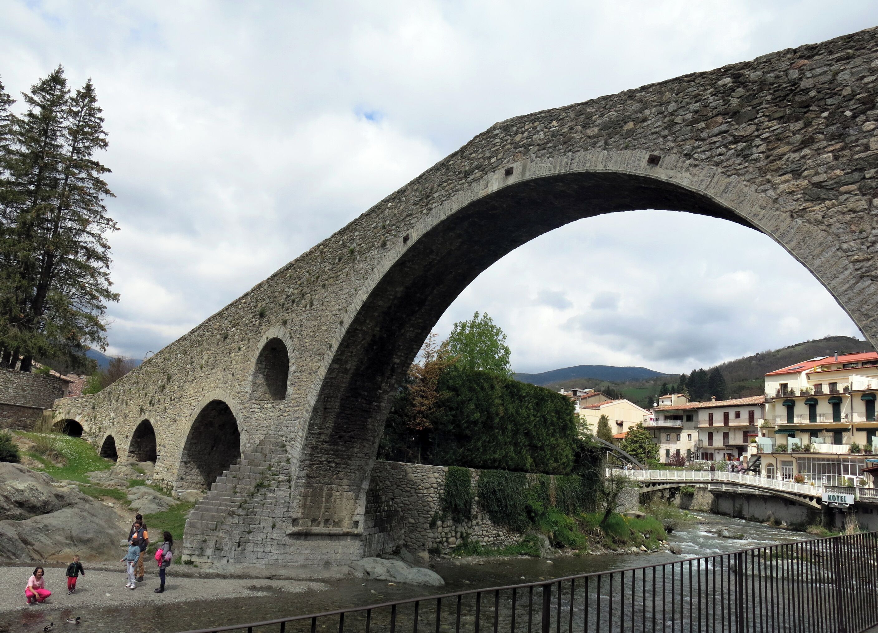 A stone bridge with multiple arches over a river, surrounded by greenery and buildings.