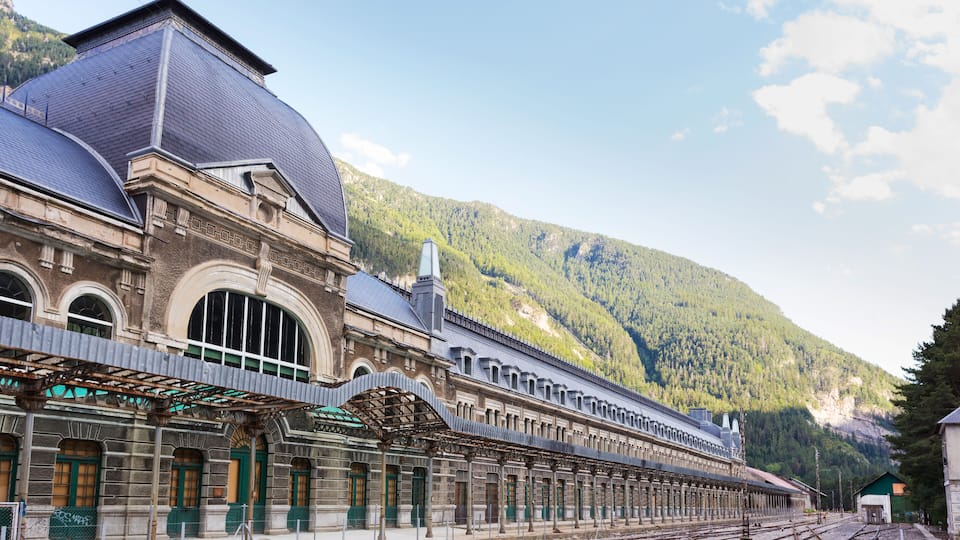 A historic train station with a large dome and arched windows, set against a mountainous backdrop.