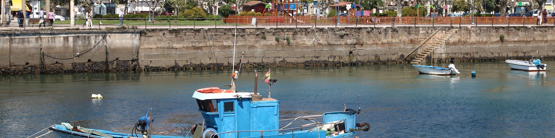A blue fishing boat docked in a calm harbor with buildings and trees in the background.
