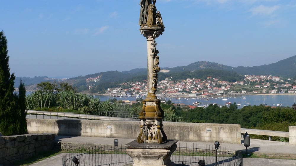 A historical monument with a statue, surrounded by a stone fence, overlooking a coastal town with boats and hills in the background.