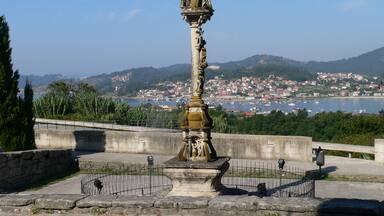 A historical monument with a statue, surrounded by a stone fence, overlooking a coastal town with boats and hills in the background.