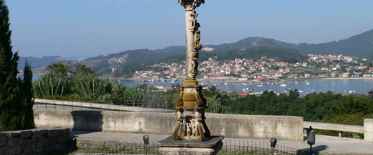 A historical monument with a statue, surrounded by a stone fence, overlooking a coastal town with boats and hills in the background.