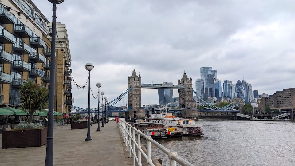 London, UK - 05.13.2024: A footpath on Butler's Wharf along outdoor seating of a restaurant and boats moored on the side of River Thames, with Tower Bridge and modern skyscrapers at the back