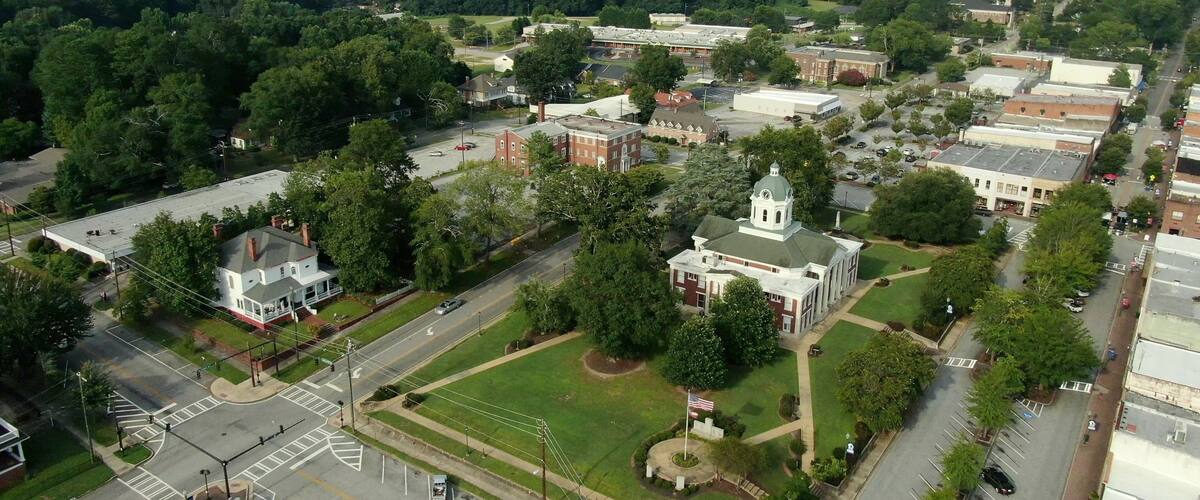 An aerial view of a town with a mix of residential and institutional buildings, surrounded by dense forests.