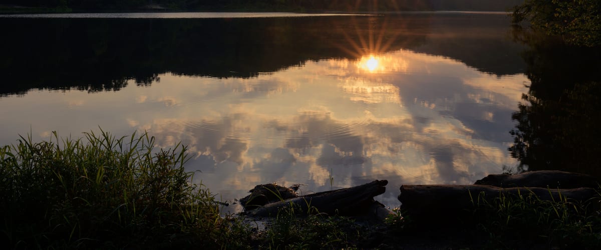 A calm lake with the sun reflecting off the water, surrounded by trees and hills.