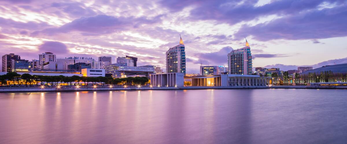 A city skyline at dusk with illuminated buildings reflecting on a calm body of water.
