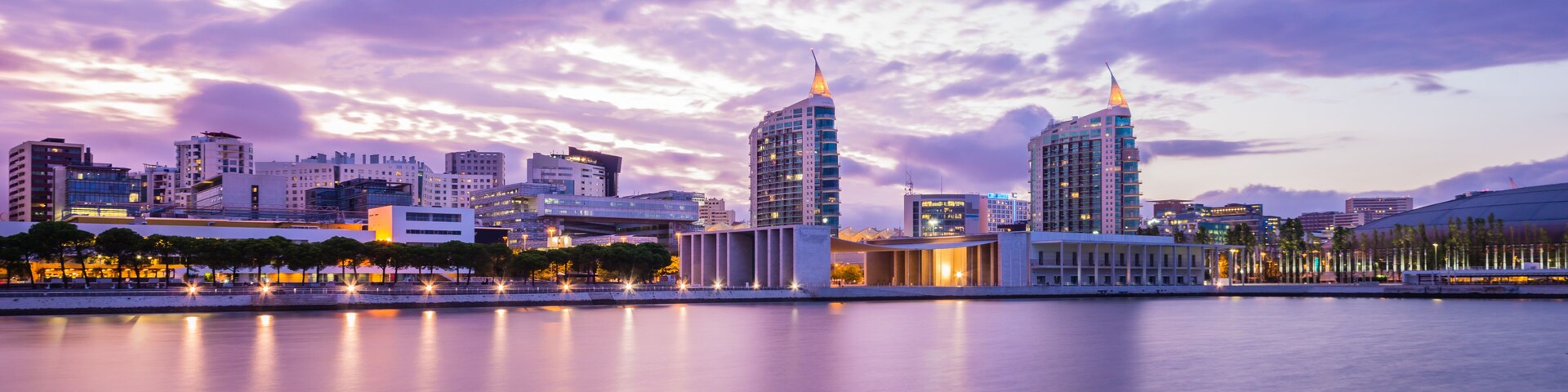 A city skyline at dusk with illuminated buildings reflecting on a calm body of water.