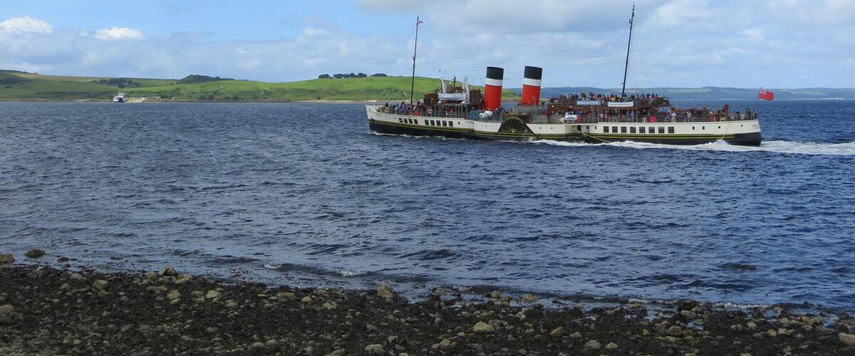 A ferry with passengers, a rocky shoreline, and a distant green landscape under a cloudy sky.