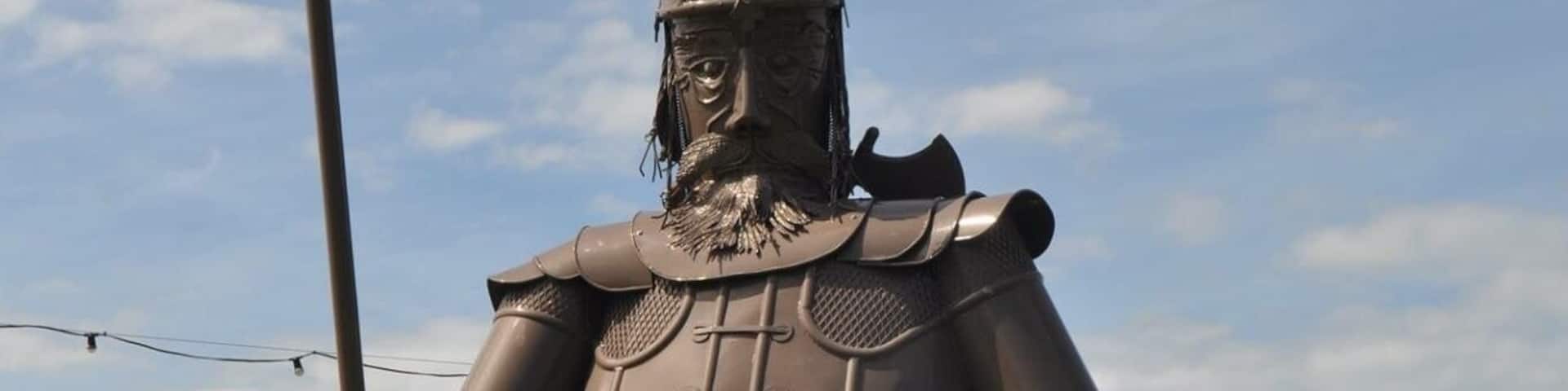 A bronze statue of a warrior holding a spear and shield, standing on a grassy area with a lake and mountains in the background.