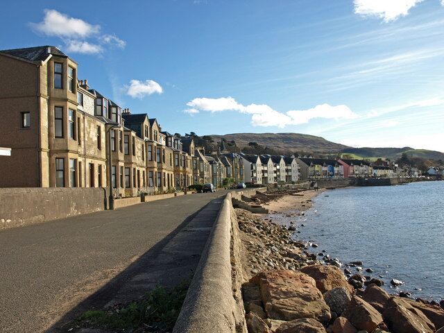 Une rue côtière bordée de maisons à étages, un rivage rocheux et un ciel dégagé.