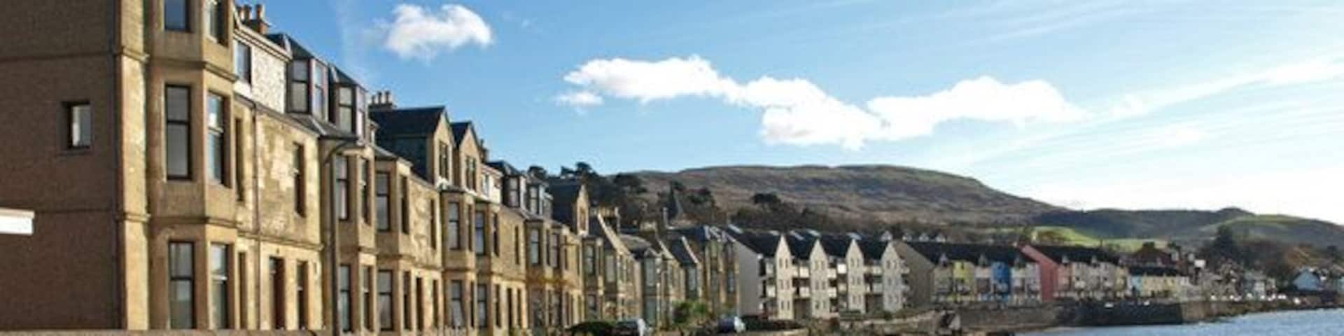 A coastal street with terraced houses, a rocky shoreline, and a clear sky.