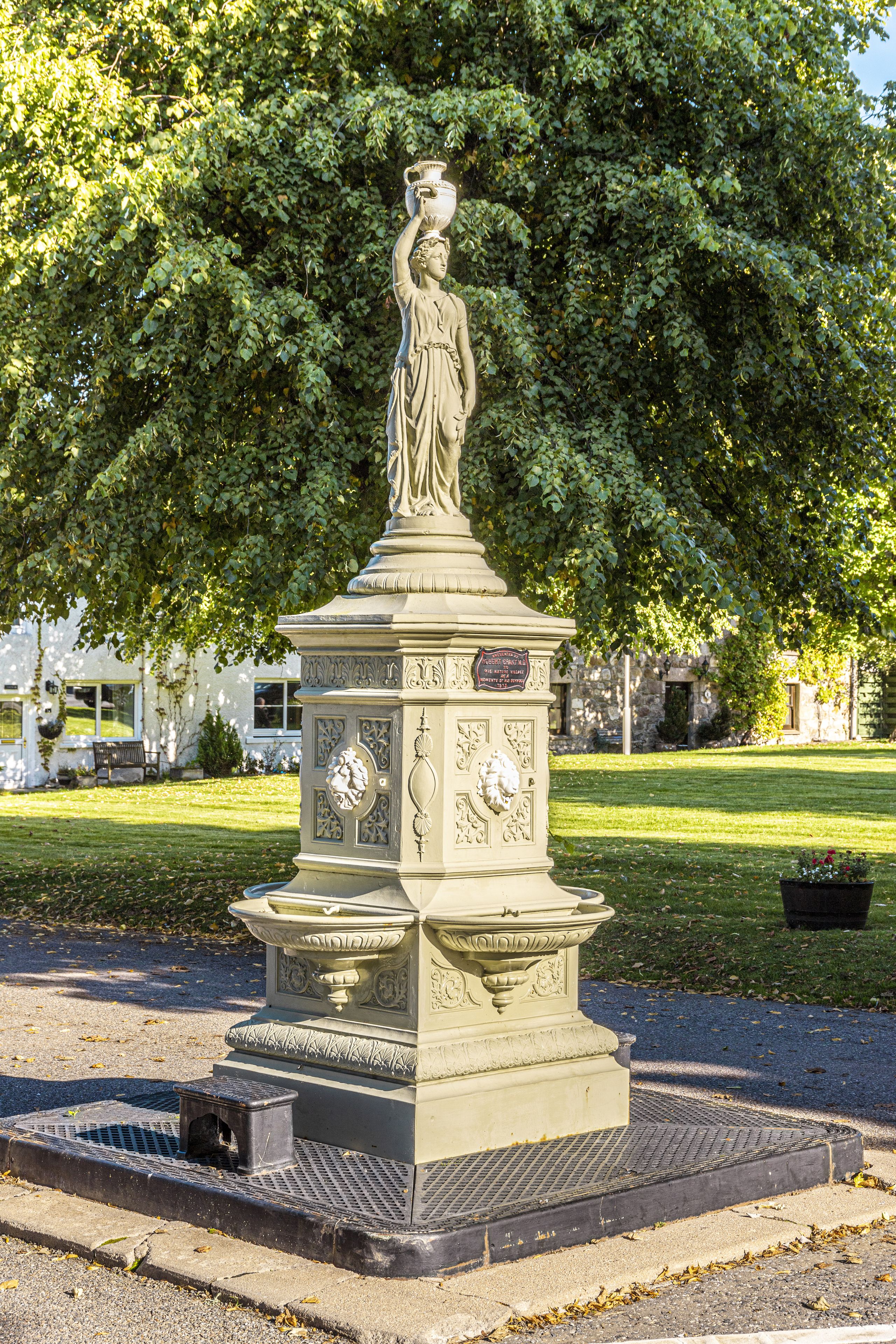 Une statue classique, posée sur un piédestal orné de sculptures complexes, se trouve dans un parc avec un grand arbre et un bâtiment en arrière-plan.