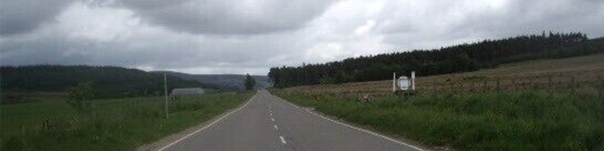 A rural road with grassy fields and a cloudy sky.