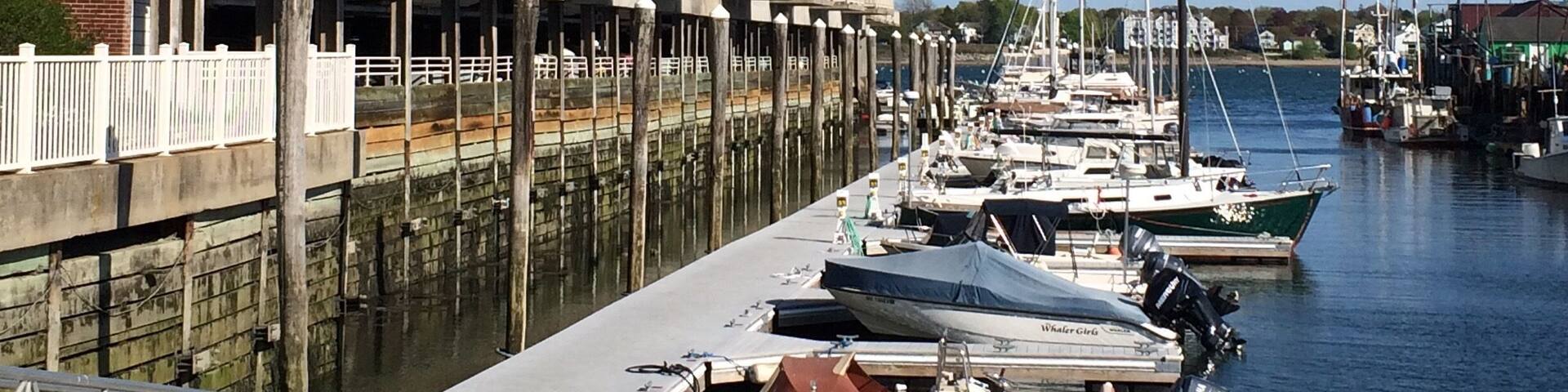 A marina with boats docked along a waterfront, a row of multi-story buildings with balconies, and a clear blue sky.