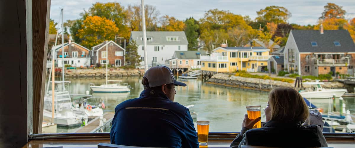 A couple sitting by a window overlooking a harbor with boats and houses.