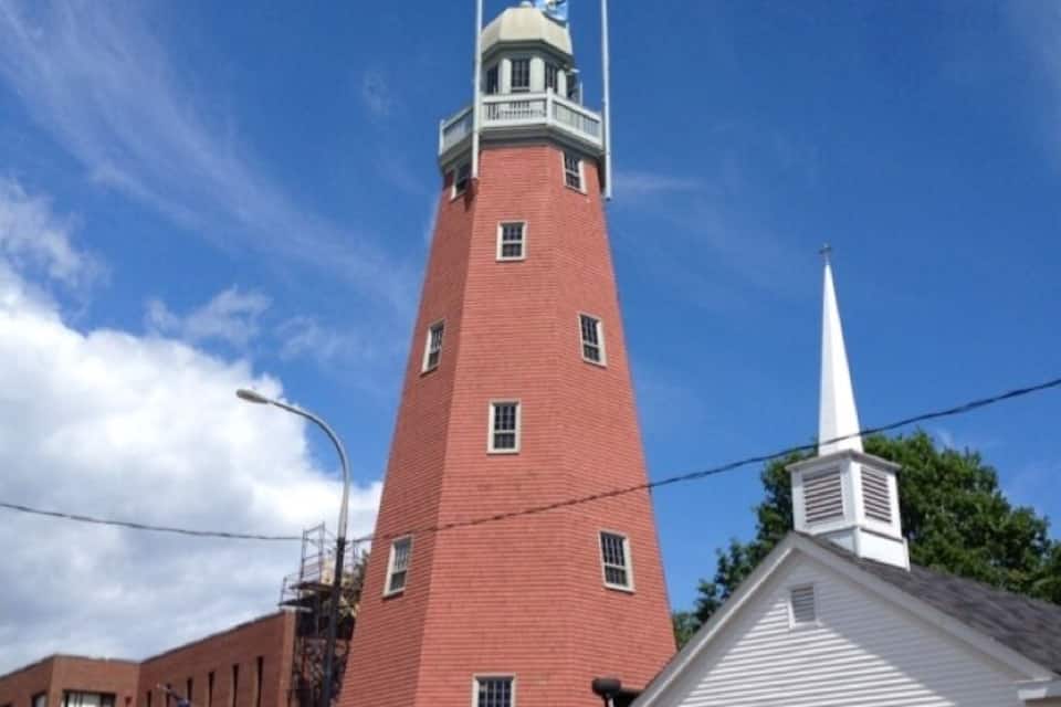 A tall brick tower with a white steeple, a small white building with a flag, and a clear blue sky.