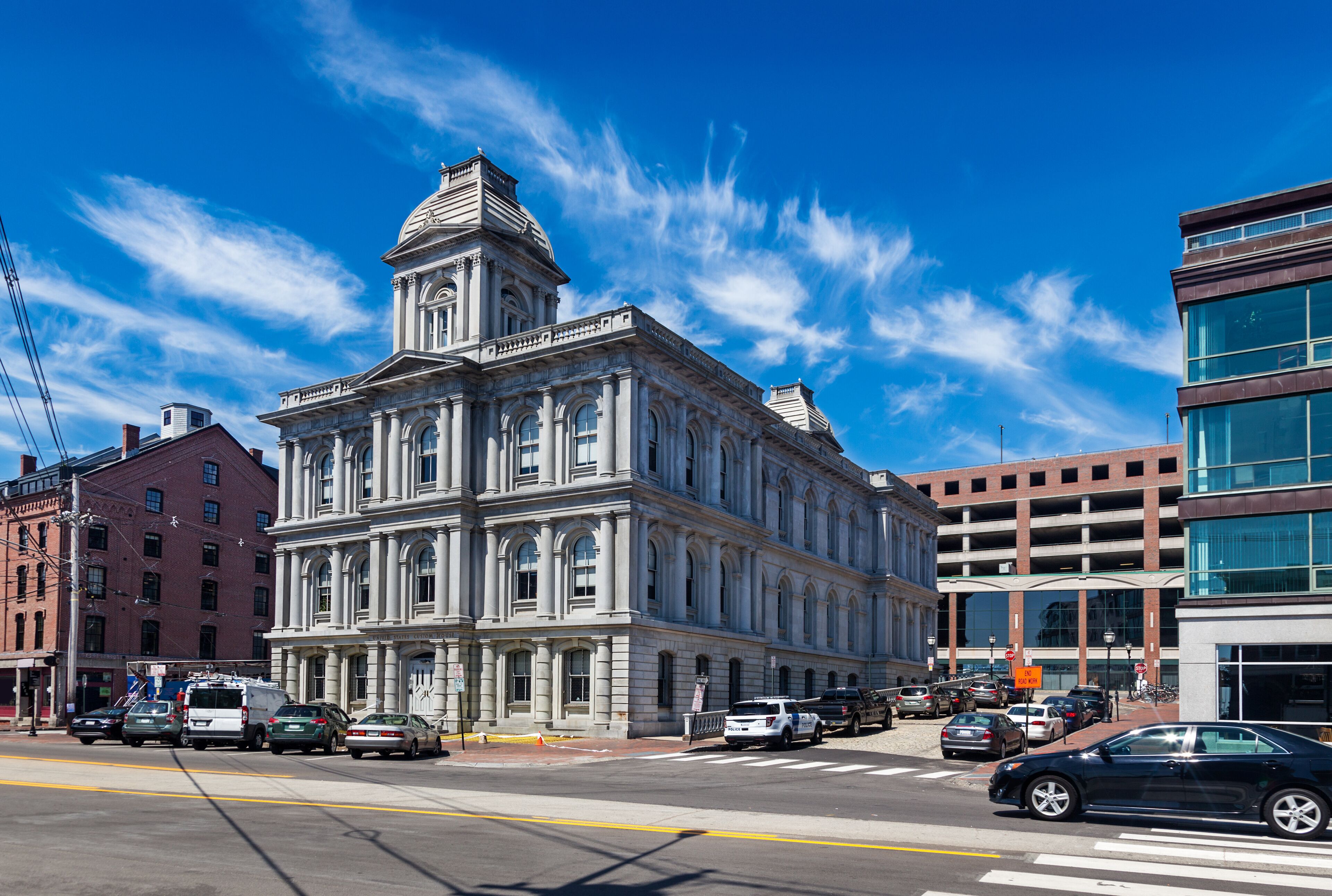 A historic building with a clock tower, surrounded by modern buildings and parked cars on a sunny day.