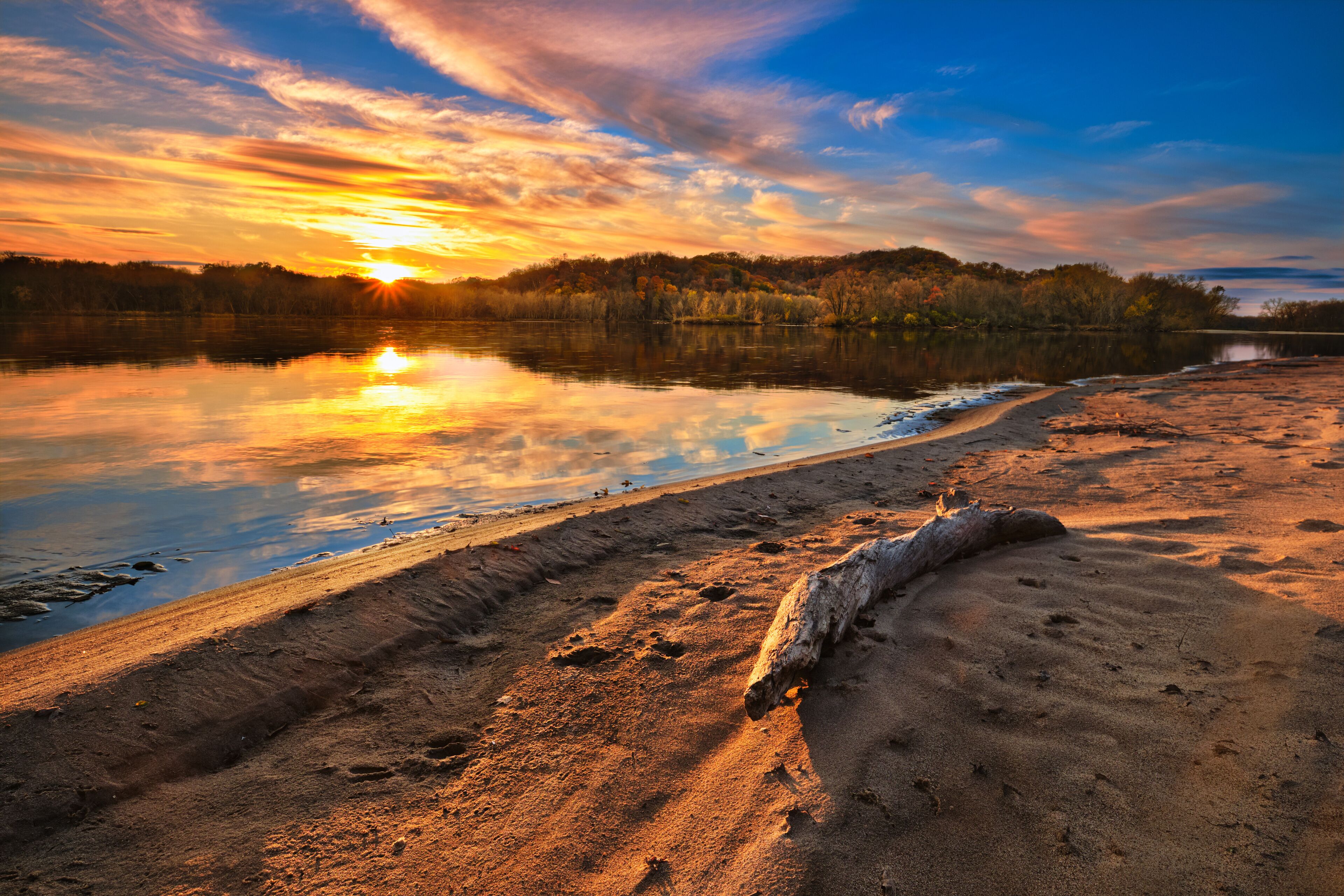 A sunset over a calm lake with a sandy shore and a fallen tree.