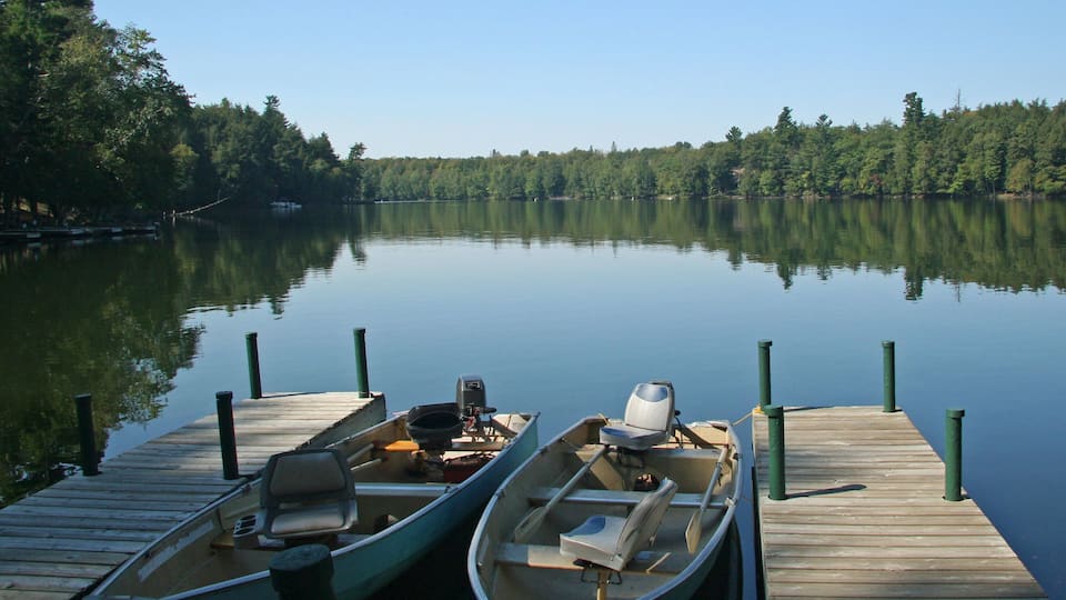 A calm lake with two boats docked at a wooden pier, surrounded by dense forest.