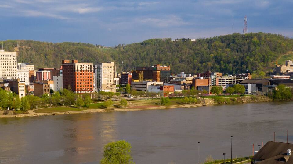 A riverside cityscape with a mix of modern and historic buildings, a bridge, and a forested hill in the background.