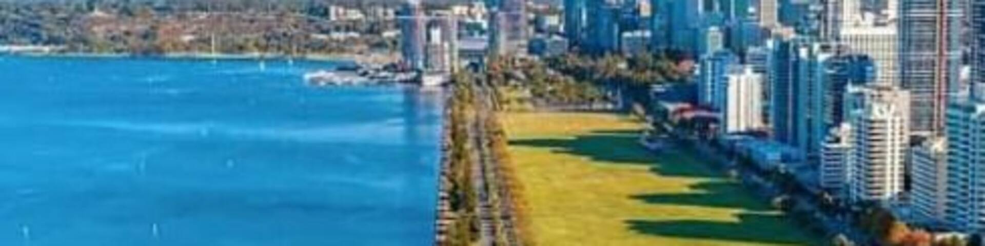 A coastal cityscape with a long promenade along the waterfront, a park, and modern high-rise buildings.