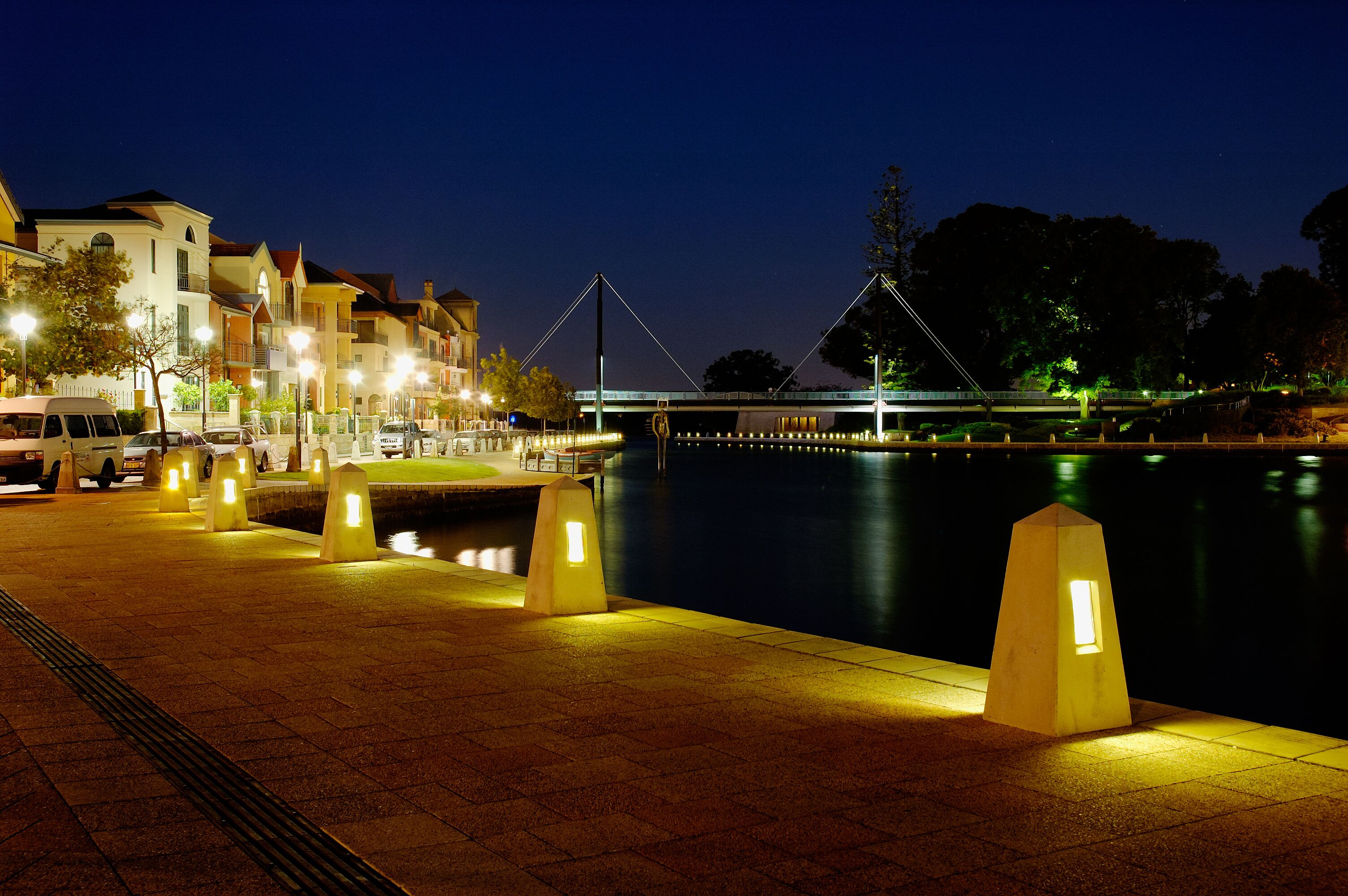 A nighttime riverside walkway with illuminated bollards, a pedestrian bridge, and buildings along the riverbank.